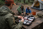 Man using a First Aid Kit Checklist while kids play in a forest campsite, showcasing essential emergency supplies.
