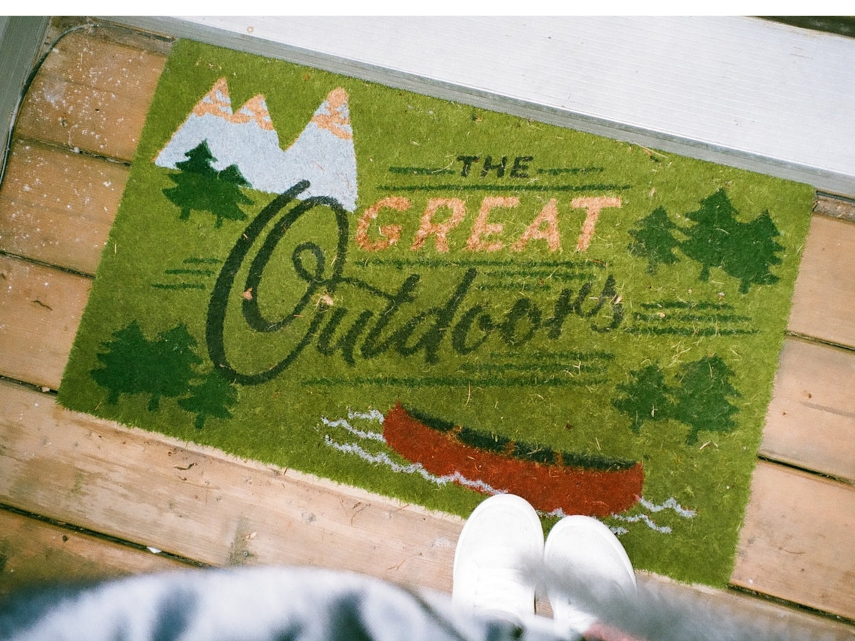 Welcome mat featuring 'THE GREAT Outdoors' design with mountains, trees, and a canoe for outdoor enthusiasts.