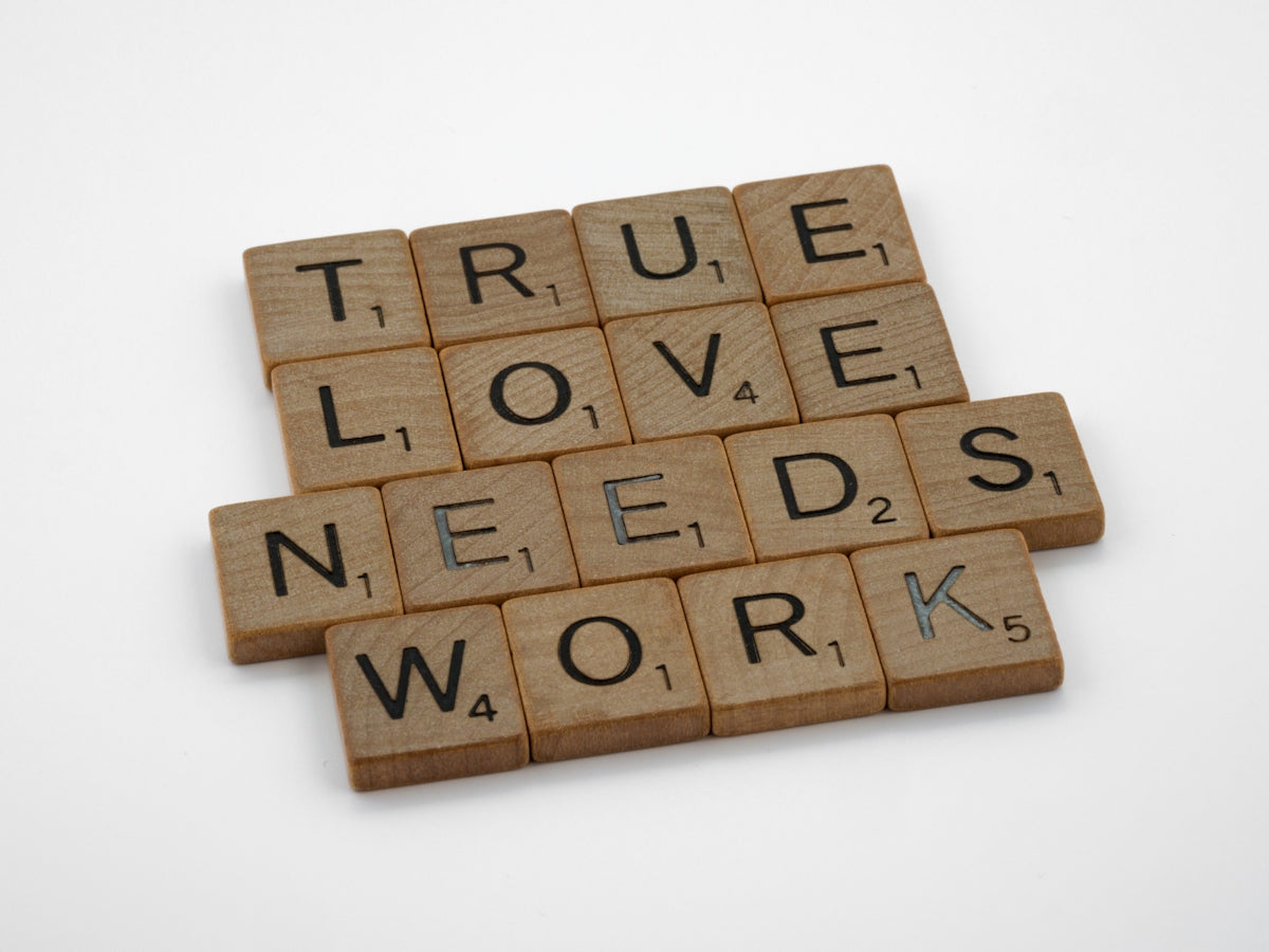Scrabble tiles spelling out 'TRUE LOVE NEEDS WORK' on a white background.