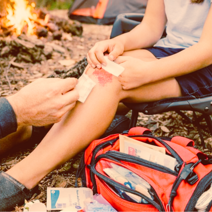 Outdoor camping scene with a person applying a bandage to another person's knee at a campsite, with a red first-aid backpack, tent, and campfire in the background.