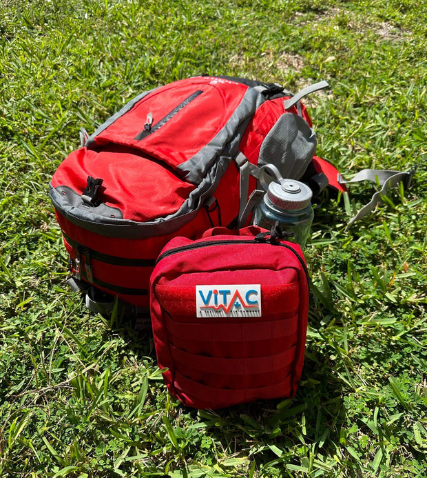 ViTAC Adventurer First Aid Kit with red backpack and water bottle on grass, ideal for outdoor activities.