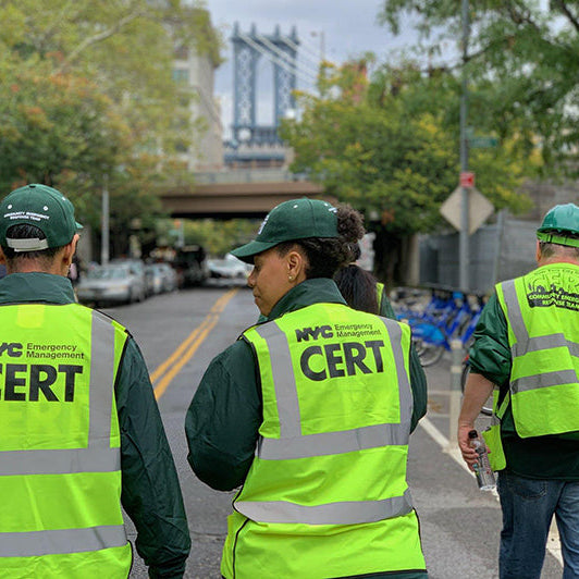 Three individuals wearing NYC CERT green vests walking on a street with trees and buildings in the background.