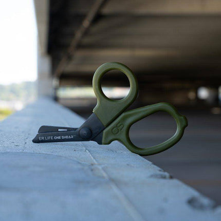MINI Compact Medical olive drab trauma shears resting on a concrete ledge with a blurred background.